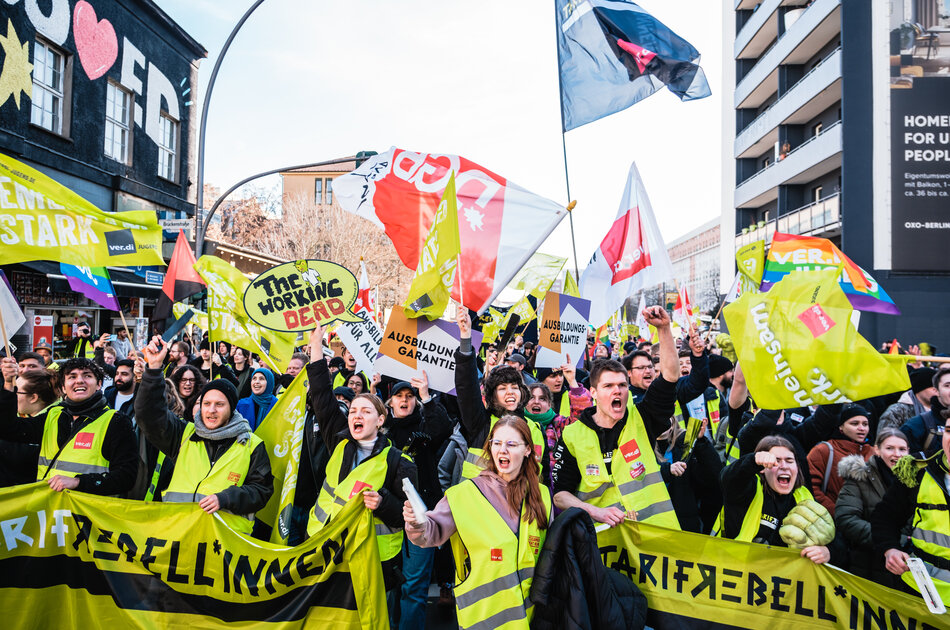 Eine Gruppe junger Streikender in gelben Westen hält Tarifrebell*innen-Banner und ruft rebellisch in die Kamera.